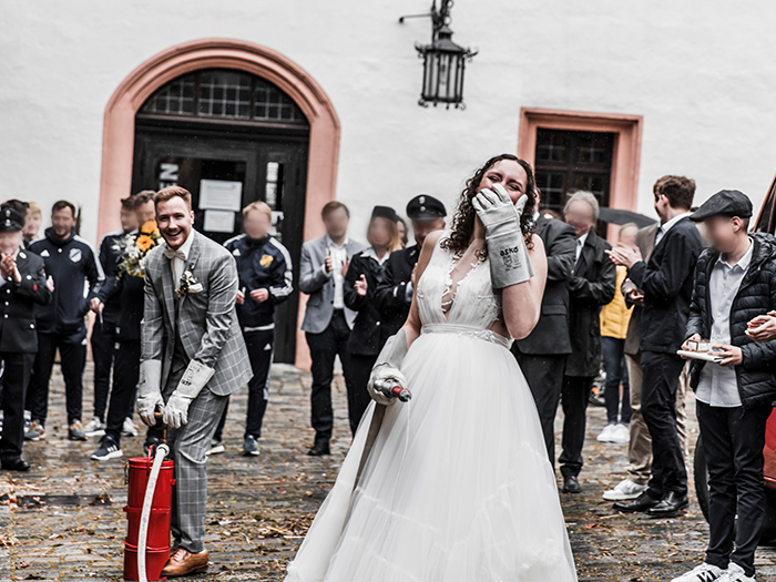 Braut spritzt mit Feuerwehrpumpe. Hält sich lachend die behandschuhte Hand vors Gesicht